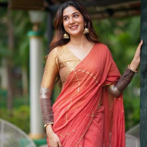 May include: A coral-colored saree featuring thin gold geometric lines, paired with a gold v-neck blouse with long brown sleeves. The ensemble includes gold jhumka earrings and a gold bracelet, showcasing a draped fabric design with metallic accents.