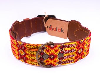 May include: A dog collar with a brown leather base and a vibrant, patterned fabric overlay in red, yellow, and orange. The collar features a metal buckle and a tag with the brand name "Aalak".