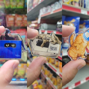May include: Three clear acrylic cat-themed keychains. The first keychain features a black cat peeking out of a blue box with the text "Lost in the City". The second keychain features a tabby cat sleeping on a white surface with the text "Lost in the City". The third keychain features an orange cat wearing a blue can on its head.