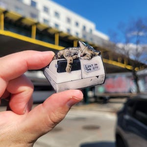 May include: A white and gray cat figurine with brown and black markings is sleeping on a white and gray box. The box has a sign that says "Lost in the City".
