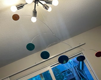 May include: A modern mobile with six circular discs in various colors, including teal, blue, red, and orange. The mobile is suspended from a silver ceiling fixture.