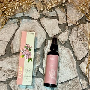 Könnte beinhalten: Zwei Flaschen mit Rosen-Duft für Raum und Wäsche. Die Flaschen sind rosa und weiß mit einem floralen Design. Der Text auf den Flaschen lautet "Rose" und "100% Natürlich".