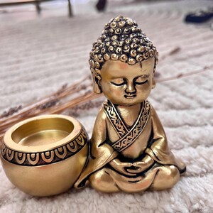 May include: A golden Buddha statue with a matching candle holder. The Buddha is in a seated meditation pose, with closed eyes and intricate details on the head and robe. The candle holder is round with a decorative pattern.