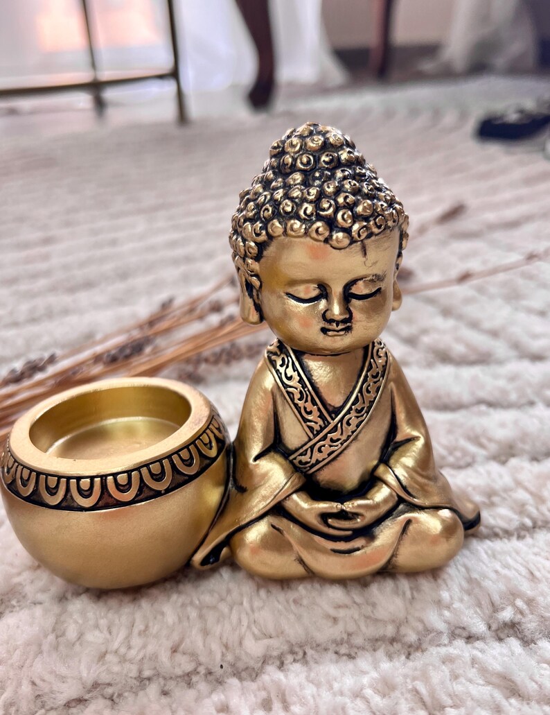May include: A golden Buddha statue with a matching candle holder. The Buddha is in a seated meditation pose, with closed eyes and intricate details on the head and robe. The candle holder is round with a decorative pattern.