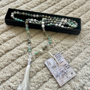 Puede incluir: Un collar de mala con cuentas blancas, verdes y moteadas, presentado en una caja negra. El collar tiene una borla blanca y una etiqueta con texto. Las cuentas están hechas de varias piedras.