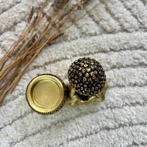 May include: A small, gold-colored Buddha statue with a textured head and a matching candle holder. The candle holder is round with an ornate border. Dried plants are in the background.