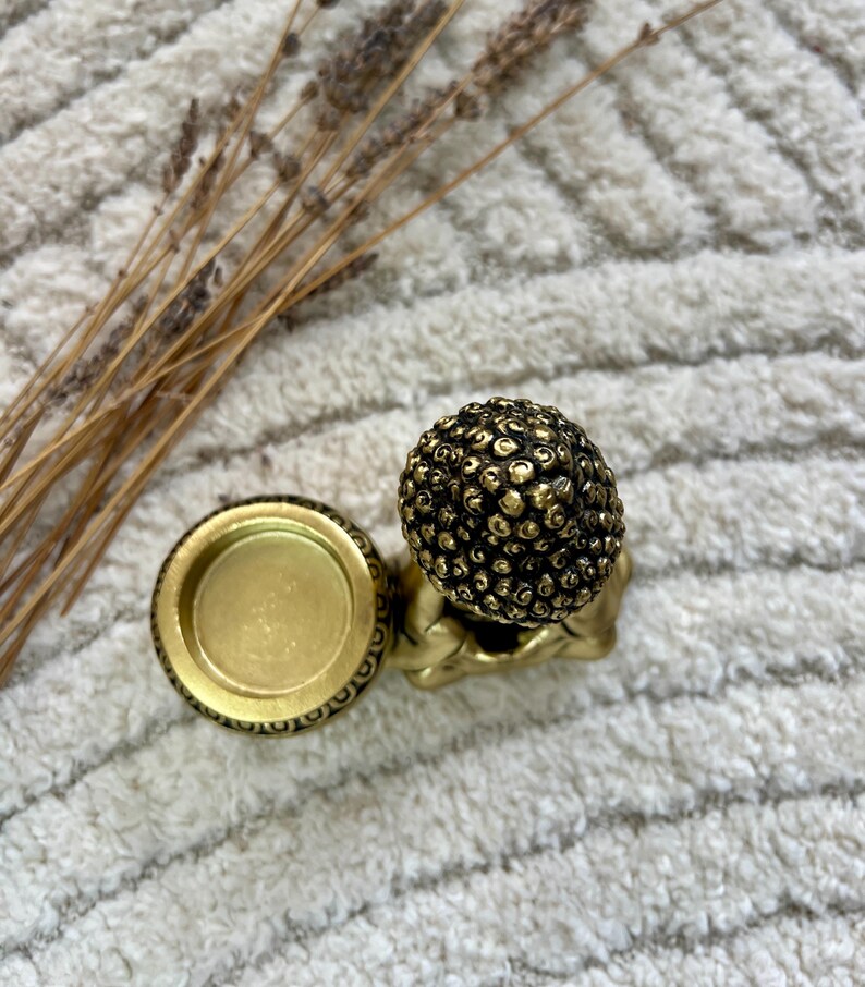 May include: A small, gold-colored Buddha statue with a textured head and a matching candle holder. The candle holder is round with an ornate border. Dried plants are in the background.