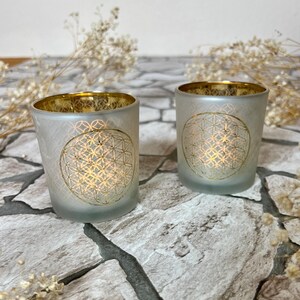 May include: Two frosted glass candle holders with a gold rim and a gold flower of life design. The candle holders are sitting on a stone surface.
