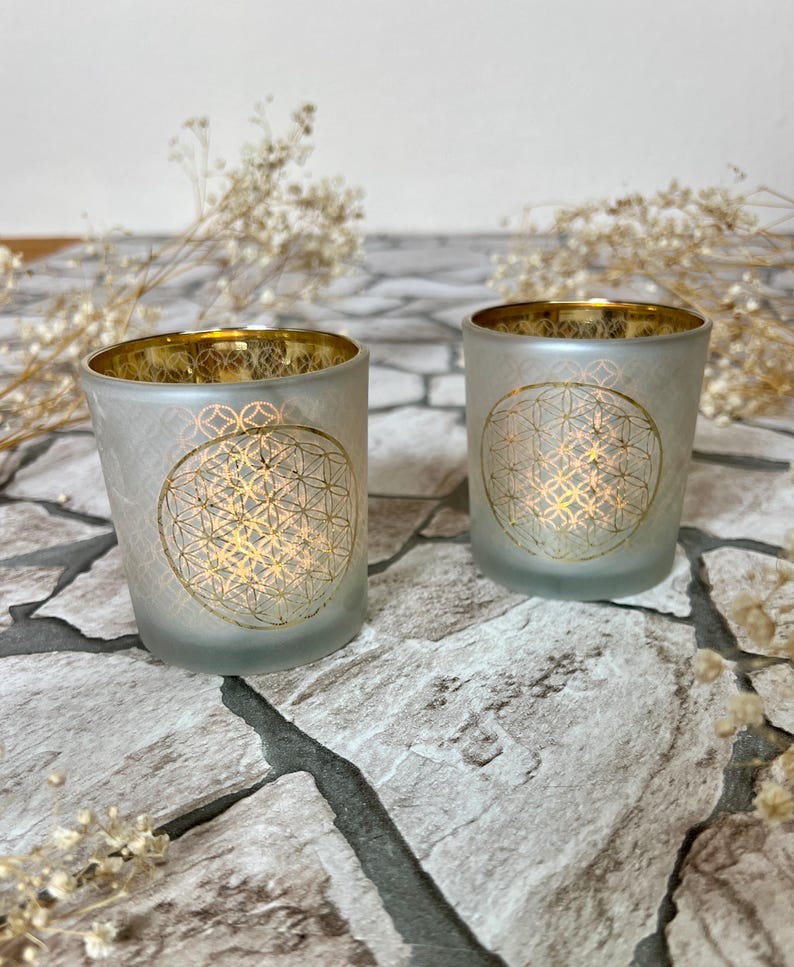May include: Two frosted glass candle holders with a gold rim and a gold flower of life design. The candle holders are sitting on a stone surface.