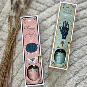 Könnte beinhalten: Zwei dekorative Raumduftboxen. Eine ist rosa mit einem Kessel- und Schmetterlingsdesign, beschriftet mit "Love Potion". Die andere ist türkis mit einem Hand- und Augendesign, beschriftet mit "Evil Eye Protection". Beide haben goldene Akzente.