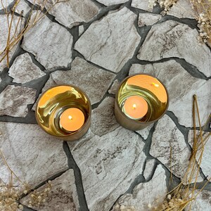 May include: Two gold candle holders with a white candle inside each. The candle holders are sitting on a gray stone surface.