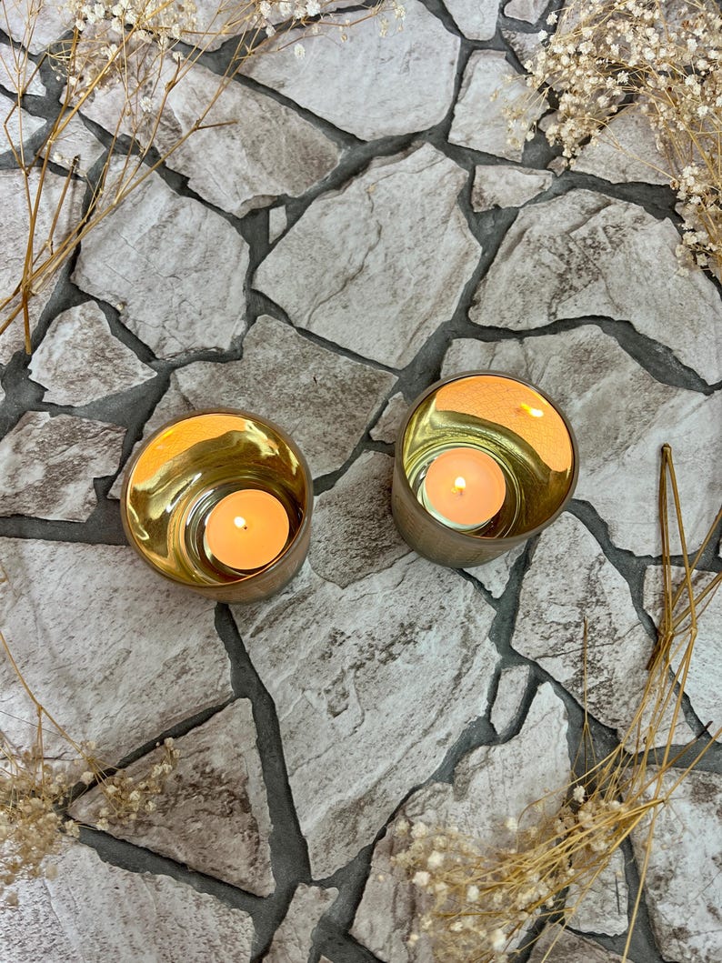 May include: Two gold candle holders with a white candle inside each. The candle holders are sitting on a gray stone surface.