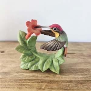 May include: A detailed figurine of a hummingbird with a red head, green body, and brown wings, reaching for a small orange flower. The bird is perched on a green leafy base, creating a nature-inspired decorative piece.