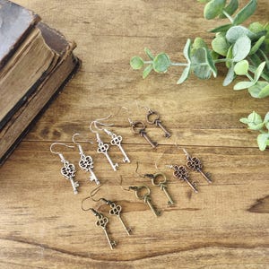May include: A collection of key-shaped earrings in various metallic finishes, including silver, bronze, and gold, displayed on a wooden surface. The earrings have intricate details and are designed with a vintage aesthetic. Green foliage and a book are in the background.