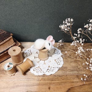 May include: A handcrafted white felt mouse with pink ears sits atop a wooden spool, placed on a white lace doily. Several other wooden spools and dried white flowers are arranged on a wooden surface, creating a rustic, vintage aesthetic.