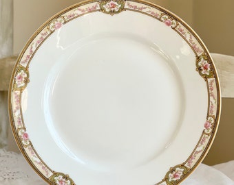 May include: A white porcelain dinner plate with a gold and green floral border. The border features pink roses and green leaves.
