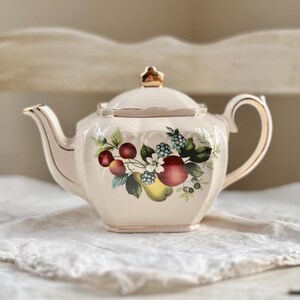 May include: A white teapot with a gold trim and a fruit design. The teapot has a lid with a gold knob and a spout on the side. The fruit design features apples, pears, cherries, and berries.