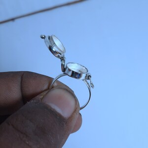 May include: A silver ring with a hinged compartment that opens to reveal an empty space. The ring is worn on a finger.