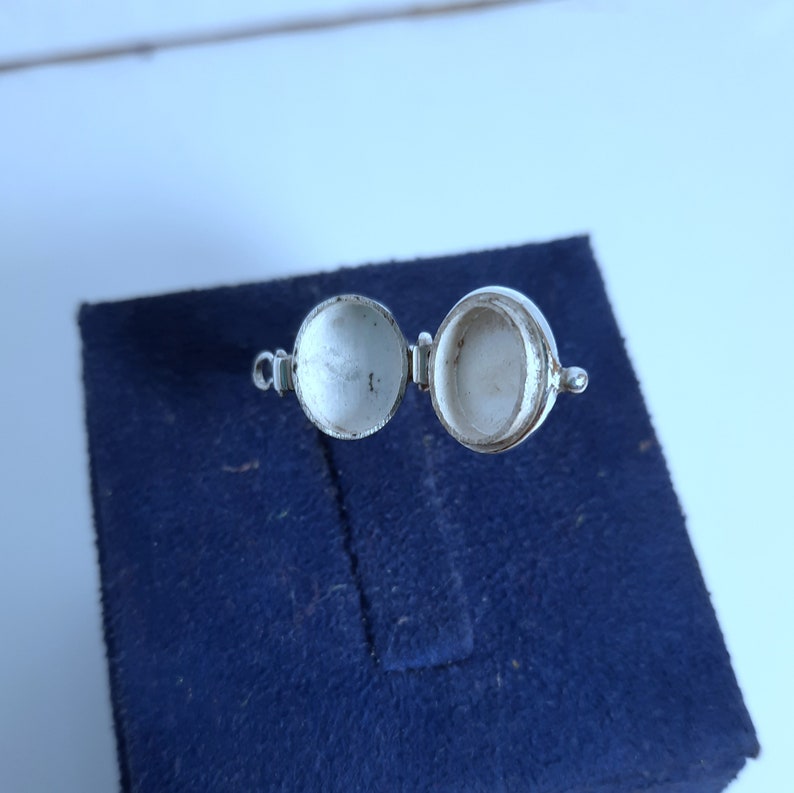 May include: A silver locket with a hinged lid, open to reveal an empty interior. The locket is on a blue velvet background.