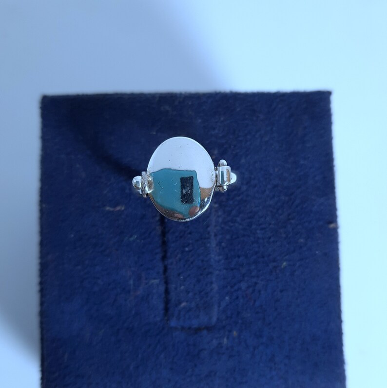May include: A silver ring with a round, smooth, polished face. The ring has a simple design with two small decorative accents on either side of the face.