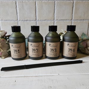 Könnte beinhalten: Vier dunkelgrüne Glasflaschen mit schwarzen Kappen und braunen Etiketten. Die Etiketten tragen den Schriftzug "Eternal" und eine Nummer von 1 bis 4. Die Flaschen sind in einer Reihe auf einer weißen Oberfläche angeordnet. Jede Flasche hat einen anderen Duft: Wacholderträume, Zitronengras, Apfelblüte und Weißer Tee.