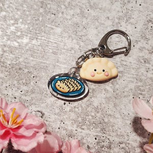 May include: A dumpling-themed keychain featuring a smiling, light-yellow dumpling charm with pink cheeks and a clear acrylic charm with a plate of dumplings. The keychain has a silver clasp.