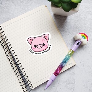 May include: A pink pig sticker with the text "stop procrastinating" sits on a lined notebook page. A pen with a rainbow and cloud topper and a glitter barrel is also on the page. A small plant in a white pot is in the background.