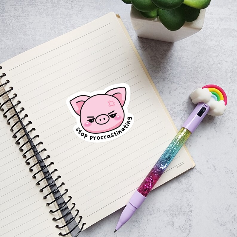 May include: A pink pig sticker with the text "stop procrastinating" sits on a lined notebook page. A pen with a rainbow and cloud topper and a glitter barrel is also on the page. A small plant in a white pot is in the background.