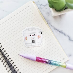 May include: A white rice cooker with a cute cartoon face and pink flowers. The rice cooker has a black handle and a digital display.