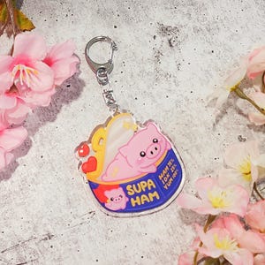 May include: A clear acrylic keychain featuring a cartoon pig in a bowl with the text "SUPA HAM." The pig is pink with a yellow and white design. The keychain has a silver clasp and is surrounded by pink flowers.