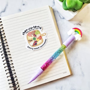 Pu&ograve; includere: Un adesivo con un tagliere di salumi disegnato in stile cartoon con il testo "char-cu-te-rie" e "fancy adult lunchables". L'adesivo &egrave; su un quaderno con una penna glitter arcobaleno.