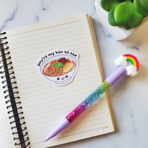 May include: A white sticker with a cartoon illustration of a bowl of bun bo hue with the text "You're my bun bo bae" on a lined notebook with a rainbow glitter pen.
