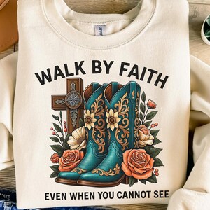 May include: Cream-colored sweatshirt with the text "WALK BY FAITH EVEN WHEN YOU CANNOT SEE." The design features teal cowboy boots, a cross, and floral accents in orange and yellow. A western-themed graphic.