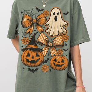 Pode incluir: T-shirt verde-oliva com um gr&aacute;fico de Halloween. O design inclui la&ccedil;os laranja, ab&oacute;boras, um fantasma, morcegos e flores. A camisa tem um ajuste folgado e mangas curtas.