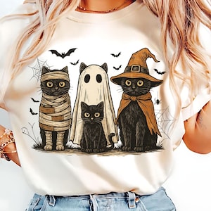 May include: A cream-colored t-shirt featuring a Halloween-themed graphic. The design includes four cartoon cats dressed as a mummy, a ghost, and a witch, with bats, spiderwebs, and spiders. The cats are in a row, with a black cat in the center.