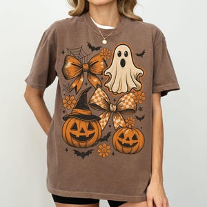 Pode incluir: Camiseta marrom com um gr&aacute;fico tem&aacute;tico de Halloween. O design inclui um fantasma, ab&oacute;boras, la&ccedil;os, flores e morcegos. A camiseta tem gola redonda e mangas curtas.