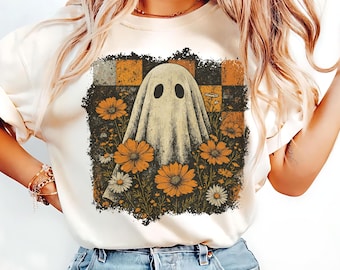 May include: Cream-colored t-shirt featuring a ghost design. The ghost is draped in a sheet and surrounded by orange and white daisies. The background has a checkered pattern in shades of brown and orange. A unique Halloween-themed graphic tee.