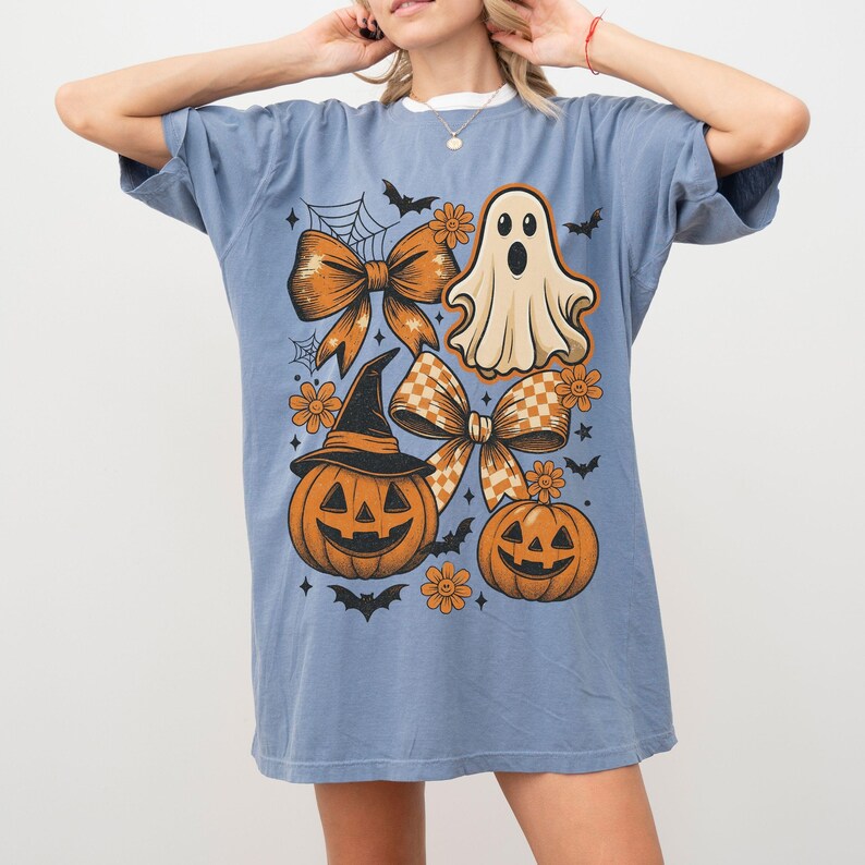 Pode incluir: Camiseta azul com um design de Halloween. O design inclui um fantasma, ab&oacute;boras, la&ccedil;os, morcegos e flores. As ab&oacute;boras t&ecirc;m rostos esculpidos e usam chap&eacute;us de bruxa. Os la&ccedil;os t&ecirc;m um padr&atilde;o xadrez.