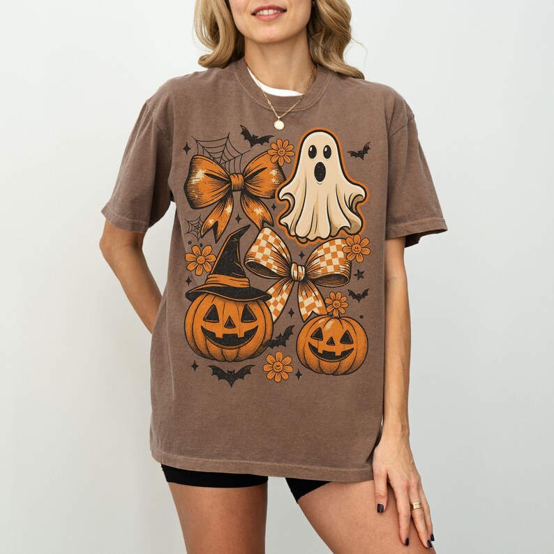 Pode incluir: Camiseta marrom com um gr&aacute;fico tem&aacute;tico de Halloween. O design inclui um fantasma, ab&oacute;boras, la&ccedil;os, flores e morcegos. A camiseta tem gola redonda e mangas curtas.