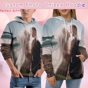 May include: A unisex hoodie with a photo of a couple on a beach. The hoodie is light blue with a white hood and a kangaroo pocket. The photo shows a couple in wedding attire, standing on a beach with a blue ocean in the background. The text "Custom Photo Unisex Hoodie" and "Perfect Gift!" is at the top of the image.