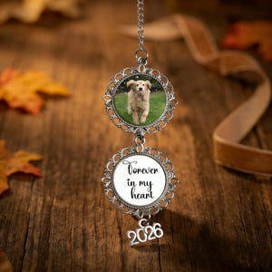 Può includere: Un ornamento color argento con una foto di un cane golden retriever in una cornice decorativa. Sotto, una seconda cornice recita "Forever in my heart" con l'anno 2026 appeso sotto. L'ornamento è appeso a una catena.