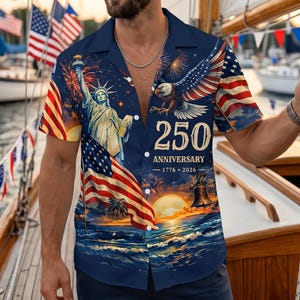 Puede incluir: Una camisa de manga corta azul marino con un estampado gráfico de la Estatua de la Libertad, un águila calva y la bandera estadounidense. El texto dice 250 ANNIVERSARY 1776 2026, junto con fuegos artificiales, un atardecer y una campana.