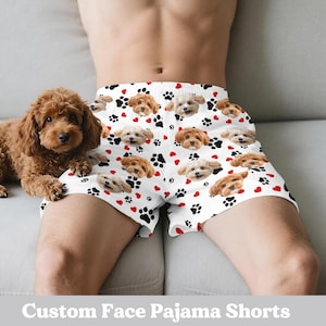 May include: White pajama shorts featuring a pattern of dog faces, black paw prints, and red hearts. The shorts have a drawstring waist. A brown dog sits beside the shorts. The text "Custom Face Pajama Shorts" is at the bottom.