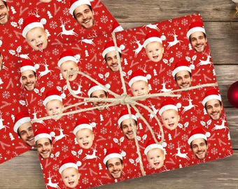 May include: Red wrapping paper featuring a repeating pattern of white reindeer, snowflakes, and holly leaves, interspersed with photographic portraits of a smiling child and a smiling adult, both wearing Santa hats, tied with natural jute twine.