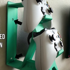 K&ouml;nnte beinhalten: Ein wandmontierter, t&uuml;rkisfarbener, 3D-gedruckter St&auml;nder f&uuml;r zwei wei&szlig;e Videospiel-Controller. Der St&auml;nder hat ein Loch zum Anbringen an einer Wand.