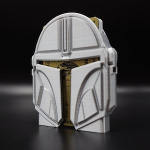May include: A white plastic card holder shaped like a Mandalorian helmet. The card holder is open and has a gold credit card inside.