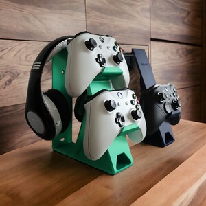 K&ouml;nnte beinhalten: Ein gr&uuml;ner Plastikst&auml;nder mit zwei wei&szlig;en Xbox-Controllern und einem schwarzen Kopfh&ouml;rer. Der St&auml;nder steht auf einer Holzoberfl&auml;che.