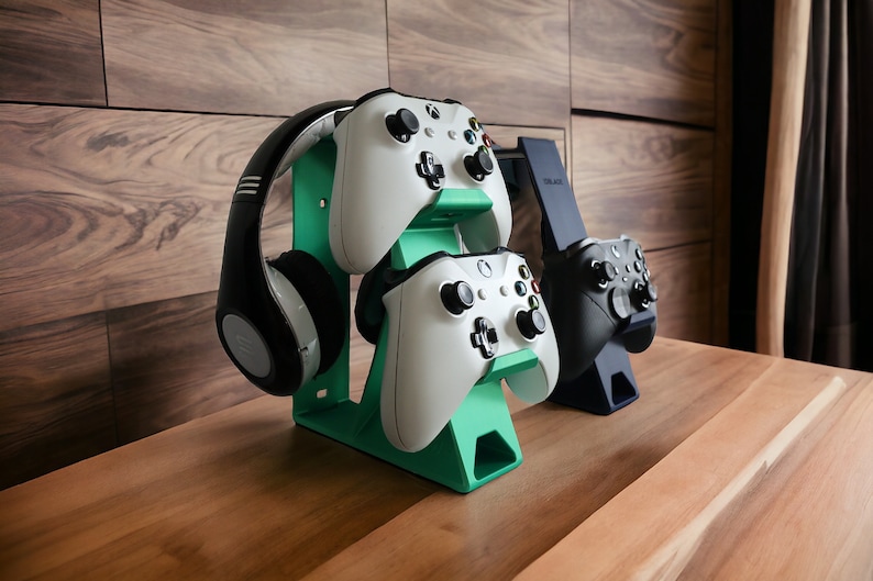 K&ouml;nnte beinhalten: Ein gr&uuml;ner Plastikst&auml;nder mit zwei wei&szlig;en Xbox-Controllern und einem schwarzen Kopfh&ouml;rer. Der St&auml;nder steht auf einer Holzoberfl&auml;che.