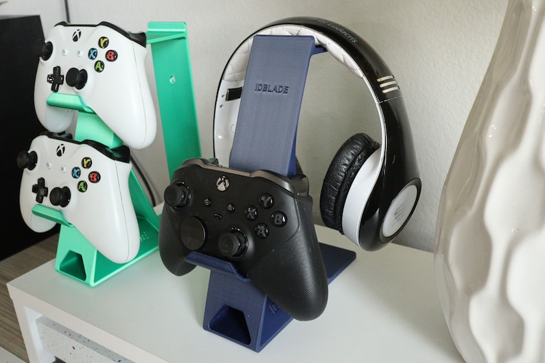 K&ouml;nnte beinhalten: Ein blauer und gr&uuml;ner 3D-gedruckter St&auml;nder h&auml;lt zwei wei&szlig;e Xbox-Controller. Ein separater blauer St&auml;nder h&auml;lt ein schwarz-wei&szlig;es Headset mit dem Text "IDBLADE" auf dem St&auml;nder.