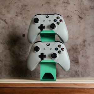 K&ouml;nnte beinhalten: Zwei wei&szlig;e Xbox-Controller werden auf einem gr&uuml;nen St&auml;nder angezeigt. Der St&auml;nder steht auf einer Holzoberfl&auml;che.